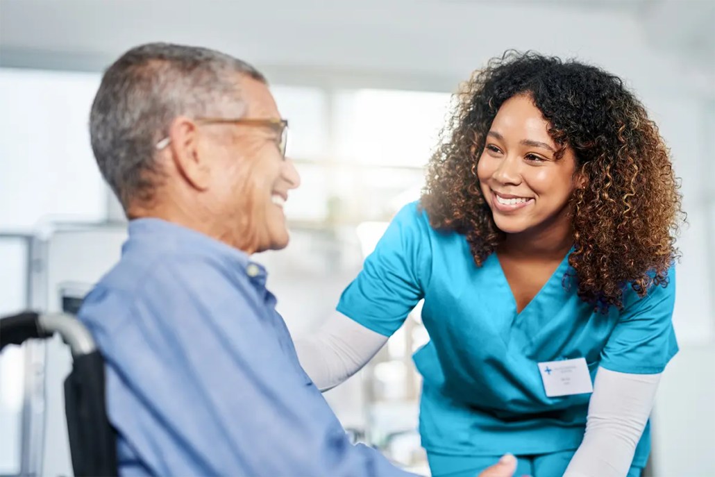 A healthcare worker in blue scrubs smiles and interacts with a senior seated in a wheelchair.