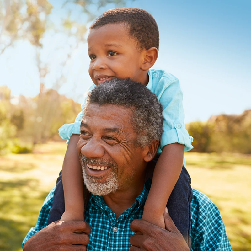 Senior man with gray hair and a beard, wearing a blue checkered shirt, smiles as he carries a young boy on his shoulders.