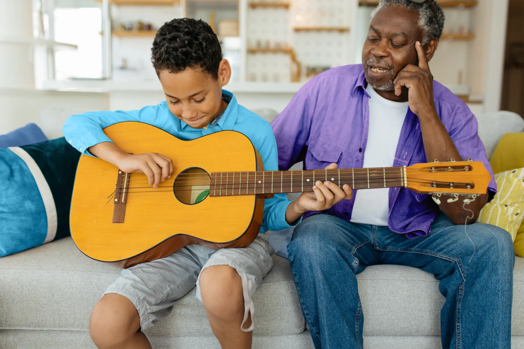 Young boy plays acoustic guitar on couch while an senior man watches proudly beside him.