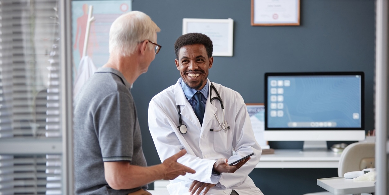 Smiling Black Doctor Holding Tablet Talking to Senior Patient in Consultation